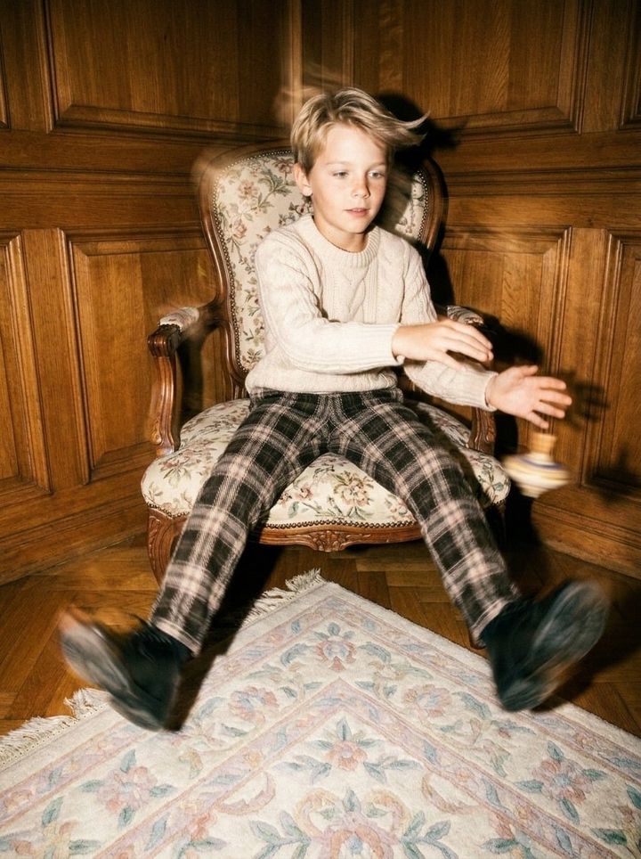 A boy in a cream knit sweater and plaid trousers in an antique armchair, legs swinging, a spinning top in motion